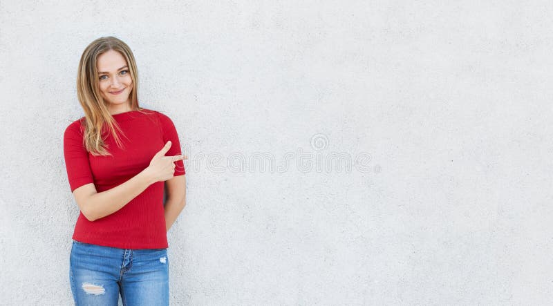 穿红色毛线衣和牛仔裤的逗人喜爱的妇女水平的画象站立在poiting与她的食指的白色混凝土墙附近在 库存照片 穿红色毛线衣和牛仔裤的逗人喜爱的妇女水平的画象站立在poiting与她的食指的白色混凝土墙附近在 库存照片