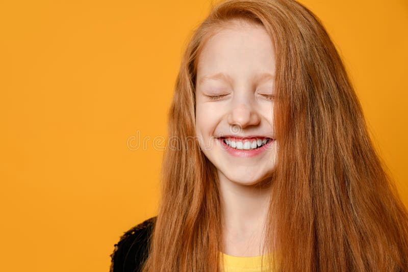 穿黑色夹克和黄色T恤的红头少女 她闭着眼睛笑着，在橙色背景上摆姿势 特写 库存照片