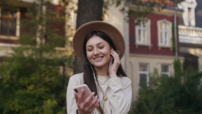 站在宅院院子里，戴着可爱帽子，微笑着的美丽现代女性，在耳机里听音乐