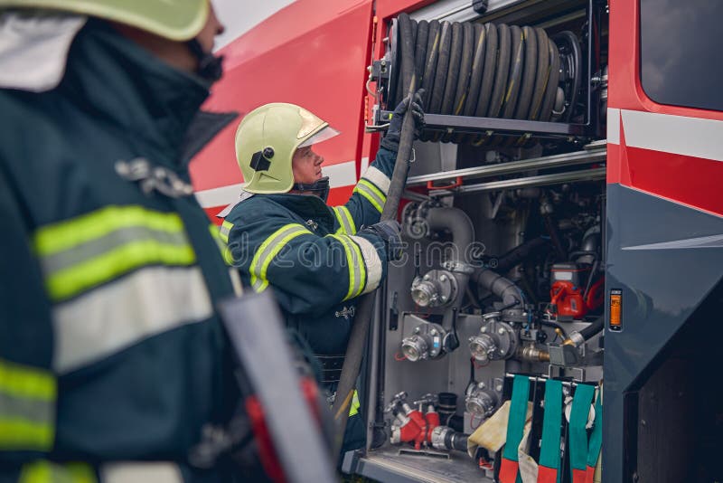 Side view of brave firefighter in protective uniform and full equipment taking hose from fire truck in order to put out the fire. Side view of brave firefighter in protective uniform and full equipment taking hose from fire truck in order to put out the fire