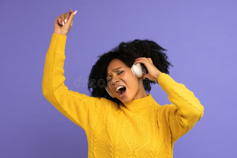 Energized biracial woman sing along, smile broadly, wears wireless headphones, has good mood, isolated on violet background. Happy dark skinned curly girl throws party, dance in rhythm of music. Energized biracial woman sing along, smile broadly, wears wireless headphones, has good mood, isolated on violet background. Happy dark skinned curly girl throws party, dance in rhythm of music