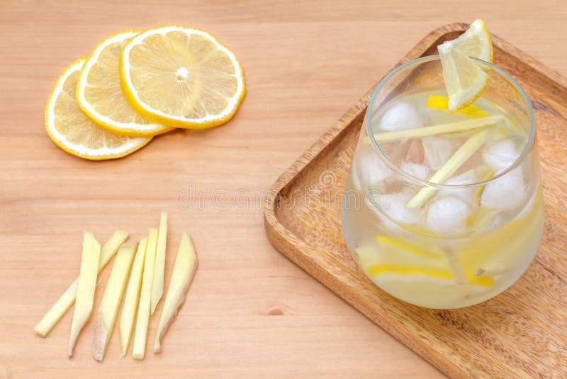 Several slices of lemon, light-brown ginger cut in pieces and a water glass full of cold refreshing lemonade with ice cubes on a wooden tray, healthy energy drink ready be served with ingredients. Several slices of lemon, light-brown ginger cut in pieces and a water glass full of cold refreshing lemonade with ice cubes on a wooden tray, healthy energy drink ready be served with ingredients