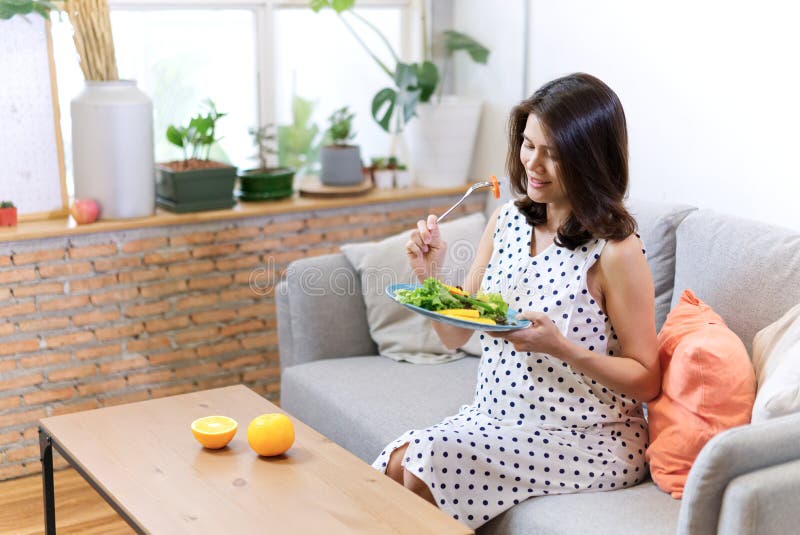 美丽的亚裔孕妇坐沙发食用沙拉有些桔子在桌上把放的她的早餐 ?? 库存图片 美丽的亚裔孕妇坐沙发食用沙拉有些桔子在桌上把放的她的早餐 ?? 库存图片