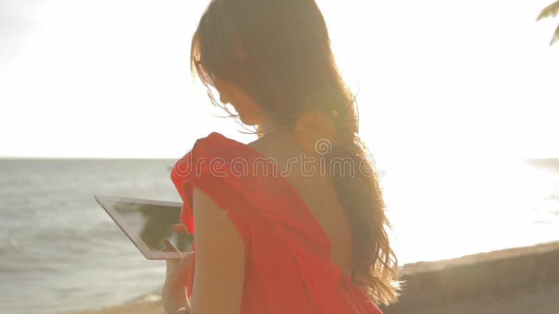美丽的女孩在一个晴朗的海滩走，使用片剂并且微笑着