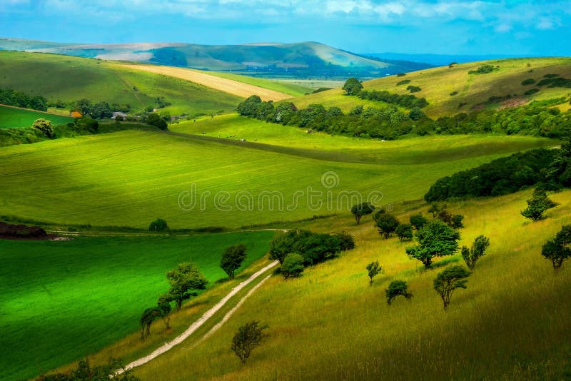 Rolling english countryside, hills on each side of the image with a dirt track running through the bottom of the hills in the middle of the image, farmland land with small trees and bushes edging rural the fields Sussex Uk beautiful painterly effect. Rolling english countryside, hills on each side of the image with a dirt track running through the bottom of the hills in the middle of the image, farmland land with small trees and bushes edging rural the fields Sussex Uk beautiful painterly effect.