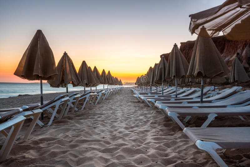 Beautiful sunset in Algarve Portugal. Beach and cliffs with closed sun umbrellas and sunbeds. Perspective on beach. Beautiful sunset in Algarve Portugal. Beach and cliffs with closed sun umbrellas and sunbeds. Perspective on beach.