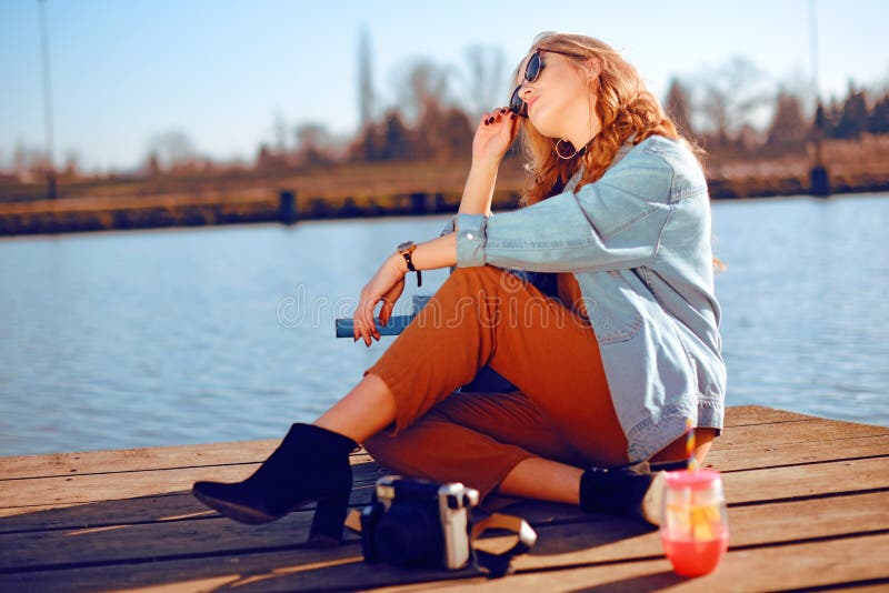 美女坐码头在河附近 在太阳镜的行家模型有白肤金发的cerly头发的 牛仔布夹克的女孩 免版税库存图片 美女坐码头在河附近 在太阳镜的行家模型有白肤金发的cerly头发的 牛仔布夹克的女孩 免版税库存图片