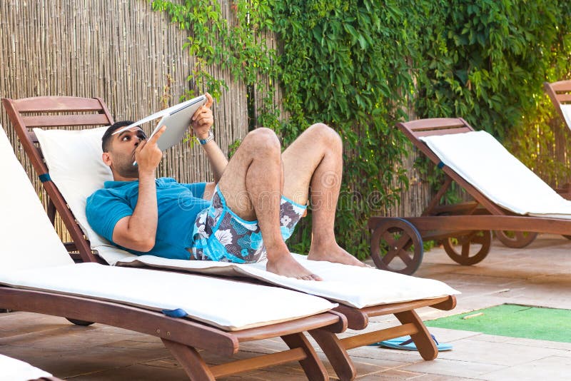 Portrait of suprised bearded young adult freelancer man in blue t-shirt and shorts lying on deckchair on poolside, holding half closed laptop and gazing trought. Lifestyle, outdoor, summer vacation. Portrait of suprised bearded young adult freelancer man in blue t-shirt and shorts lying on deckchair on poolside, holding half closed laptop and gazing trought. Lifestyle, outdoor, summer vacation