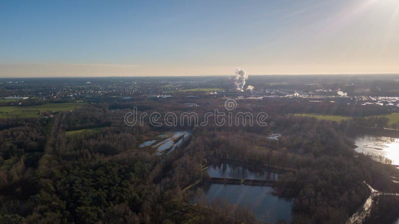 This aerial photo contrasts the tranquil beauty of a natural landscape with the distant bustle of industrial activity. In the foreground, serene bodies of water are surrounded by dense woodlands, reflecting the calm, clear sky. As the view extends, it captures the open fields, which serve as a buffer before giving way to the industrial complex. Smokestacks punctuate the horizon, releasing plumes into the air, a stark reminder of the region&#x27;s economic activity. The sunlight flares in the corner, suggesting the image was captured late in the day, highlighting the complex interplay between industry and nature. Industrial Progress Amidst Natural Serenity. High quality photo. This aerial photo contrasts the tranquil beauty of a natural landscape with the distant bustle of industrial activity. In the foreground, serene bodies of water are surrounded by dense woodlands, reflecting the calm, clear sky. As the view extends, it captures the open fields, which serve as a buffer before giving way to the industrial complex. Smokestacks punctuate the horizon, releasing plumes into the air, a stark reminder of the region&#x27;s economic activity. The sunlight flares in the corner, suggesting the image was captured late in the day, highlighting the complex interplay between industry and nature. Industrial Progress Amidst Natural Serenity. High quality photo