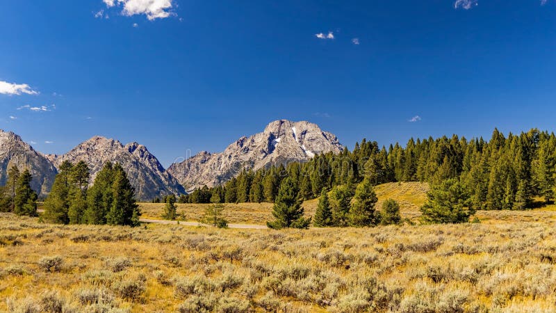 Majestic Mount Moran with its jagged peaks and small glaciers on a clear September day with a deep haze on the horizon from unchecked Canadian Forest Fires. The distinctive dark vein of igneous rock marks a period of volcanic activity from a long time ago. Mount St. John on the left leads up to Grand Teton. The slopes are covered in alpine fir and subalpine fir trees up to the harsh environment of the tree line above which only rocks occur. While the flats below and across Jackson Lake are covered in grasses and timber of Lodgepole pine and spruce varieties, the pothole region is mostly grasses and sage. Majestic Mount Moran with its jagged peaks and small glaciers on a clear September day with a deep haze on the horizon from unchecked Canadian Forest Fires. The distinctive dark vein of igneous rock marks a period of volcanic activity from a long time ago. Mount St. John on the left leads up to Grand Teton. The slopes are covered in alpine fir and subalpine fir trees up to the harsh environment of the tree line above which only rocks occur. While the flats below and across Jackson Lake are covered in grasses and timber of Lodgepole pine and spruce varieties, the pothole region is mostly grasses and sage.