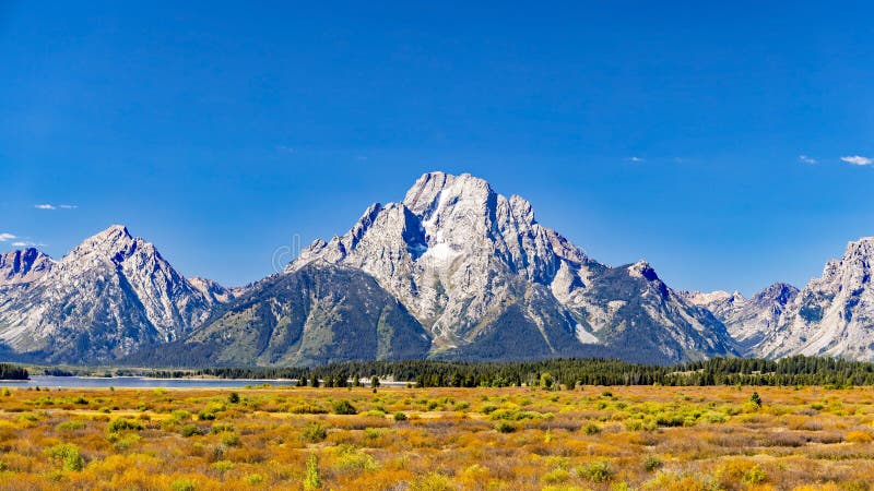 Majestic Mount Moran with its jagged peaks and small glaciers on a clear September day with a deep haze on the horizon from unchecked Canadian Forest Fires. The distinctive dark vein of igneous rock marks a period of volcanic activity from a long time ago. Mount St. John on the left leads up to Grand Teton. The slopes are covered in alpine fir and subalpine fir trees up to the harsh environment of the tree line above which only rocks occur. While the flats below and across Jackson Lake are covered in grasses and timber of Lodgepole pine and spruce varieties. Majestic Mount Moran with its jagged peaks and small glaciers on a clear September day with a deep haze on the horizon from unchecked Canadian Forest Fires. The distinctive dark vein of igneous rock marks a period of volcanic activity from a long time ago. Mount St. John on the left leads up to Grand Teton. The slopes are covered in alpine fir and subalpine fir trees up to the harsh environment of the tree line above which only rocks occur. While the flats below and across Jackson Lake are covered in grasses and timber of Lodgepole pine and spruce varieties.