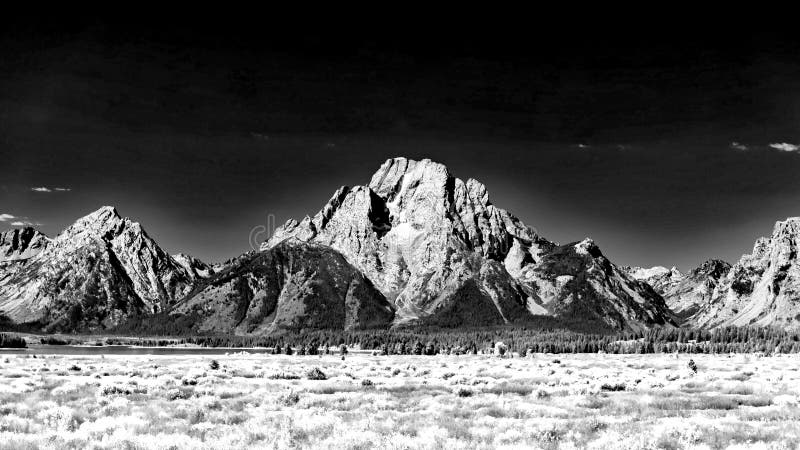 Majestic Mount Moran with its jagged peaks and small glaciers on a clear September day with a deep haze on the horizon from unchecked Canadian Forest Fires. The distinctive dark vein of igneous rock marks a period of volcanic activity from a long time ago. Mount St. John on the left leads up to Grand Teton. The slopes are covered in alpine fir and subalpine fir trees up to the harsh environment of the tree line above which only rocks occur. While the flats below and across Jackson Lake are covered in grasses and timber of Lodgepole pine and spruce varieties. Majestic Mount Moran with its jagged peaks and small glaciers on a clear September day with a deep haze on the horizon from unchecked Canadian Forest Fires. The distinctive dark vein of igneous rock marks a period of volcanic activity from a long time ago. Mount St. John on the left leads up to Grand Teton. The slopes are covered in alpine fir and subalpine fir trees up to the harsh environment of the tree line above which only rocks occur. While the flats below and across Jackson Lake are covered in grasses and timber of Lodgepole pine and spruce varieties.