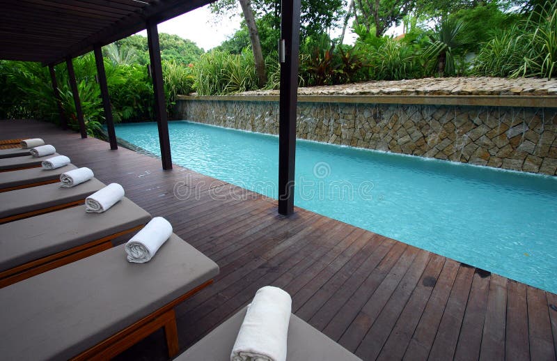 A turquoise blue resort hotel swimming pool with sheltered wooden deck on one side and poolside loungers in a row. Beach towels are rolled up and placed on chaise lounge chairs. With lush and natural tropical landscaping on other sides of the pool for exotic green forest mood. Waterfall feature on one side of pool with water cascading down artificial man made rock wall. Horizontal color format. Location is five star seaside resort and spa in Sentosa island, Singapore. Nobody in picture. A turquoise blue resort hotel swimming pool with sheltered wooden deck on one side and poolside loungers in a row. Beach towels are rolled up and placed on chaise lounge chairs. With lush and natural tropical landscaping on other sides of the pool for exotic green forest mood. Waterfall feature on one side of pool with water cascading down artificial man made rock wall. Horizontal color format. Location is five star seaside resort and spa in Sentosa island, Singapore. Nobody in picture.