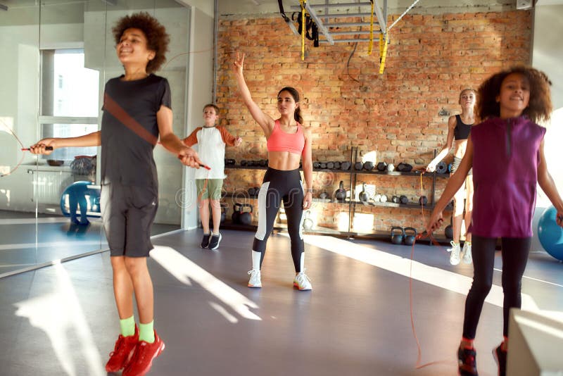 Full-length shot of female trainer controlling, looking after kids, while they are jumping rope in gym. Sport, healthy life, physical education concept. Horizontal shot. Selective focus. Full-length shot of female trainer controlling, looking after kids, while they are jumping rope in gym. Sport, healthy life, physical education concept. Horizontal shot. Selective focus