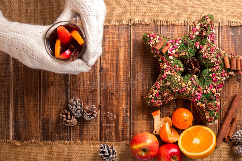 Mulled wine in woman hands in white knitted gloves near spices and fruit ingredients on wooden table. warming drink. Christmas. New year. Top view. Mulled wine in woman hands in white knitted gloves near spices and fruit ingredients on wooden table. warming drink. Christmas. New year. Top view.