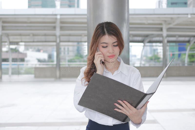 谈话在电话和在她的手上的相当年轻亚裔女商人正面图看文件外部办公室 图库摄影 谈话在电话和在她的手上的相当年轻亚裔女商人正面图看文件外部办公室 图库摄影