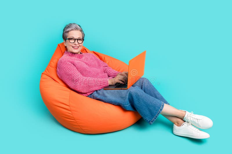 Full length photo of intelligent senior woman wear pink sweater lay on bean bag typing email on laptop isolated on teal color background. Full length photo of intelligent senior woman wear pink sweater lay on bean bag typing email on laptop isolated on teal color background.