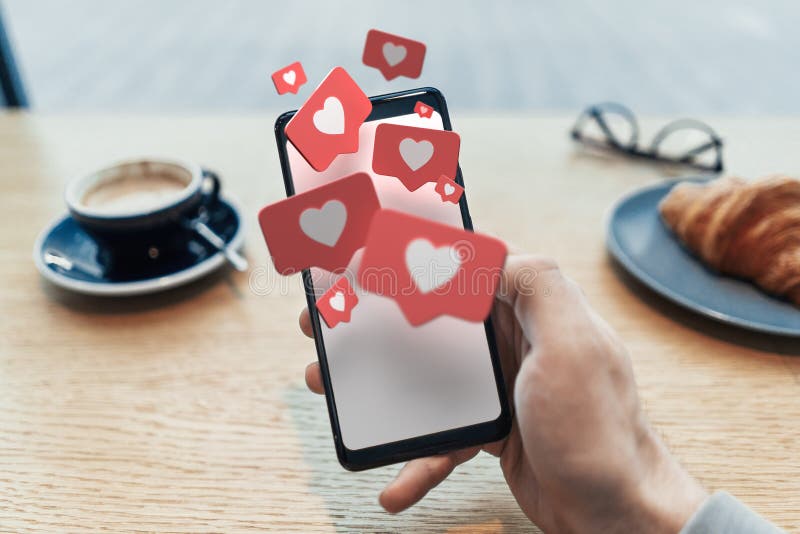 Red heart Like symbols on blank mobile phone screen. Close up of man hand holding mobile phone in cafe. 3d rendering. Social media concept. Red heart Like symbols on blank mobile phone screen. Close up of man hand holding mobile phone in cafe. 3d rendering. Social media concept.