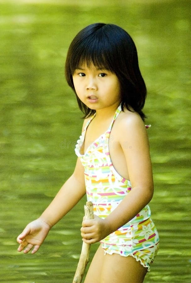 A cute young Asian girl in a swimsuit wades in a stream with a walking stick. A cute young Asian girl in a swimsuit wades in a stream with a walking stick.