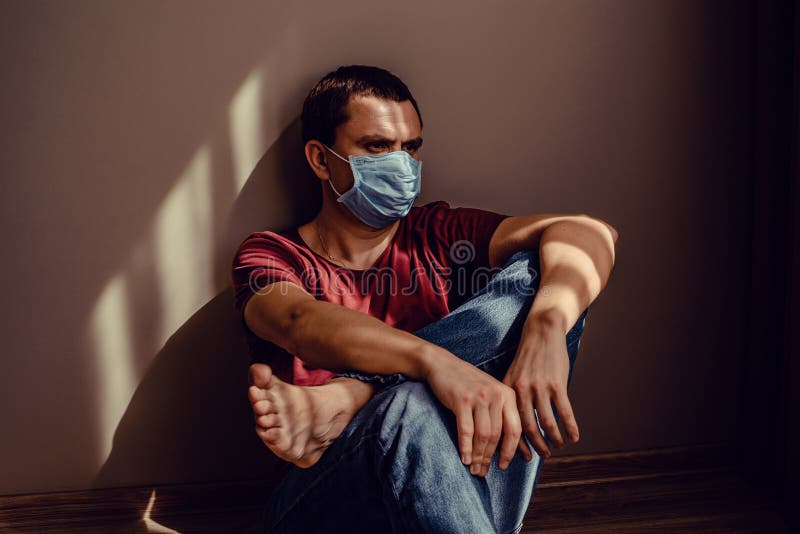 身穿医用面具的可怜成年男子坐在墙上，身上有条纹形的光影图案 保持隔离 库存图片