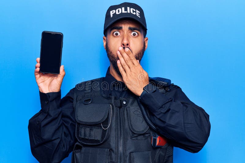 身穿警服的西班牙裔年轻人手持智能手机，嘴巴上手震惊，害怕犯错 免版税库存图片
