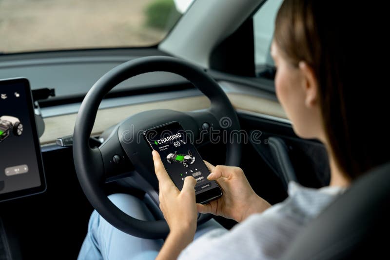 Young woman inside the vehicle, checking EV vehicle&#x27;s application on battery recharging electricity status display on smartphone screen in modern EV car on her road trip journey. Exalt. Young woman inside the vehicle, checking EV vehicle&#x27;s application on battery recharging electricity status display on smartphone screen in modern EV car on her road trip journey. Exalt