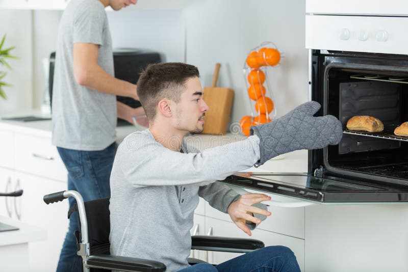年轻残疾男子坐在轮椅上打开烤箱准备食物 库存照片