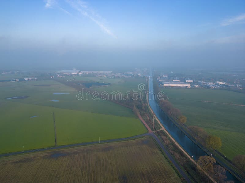 The image offers a bird&#x27;s-eye view of a serene rural landscape shrouded in mist. In the foreground, a canal cuts a clear, straight path between fields of varying shades of green, hinting at different crops or stages of cultivation. The mist creates a soft focus effect, especially on the horizon where the outlines of distant buildings and trees become less distinct. The early morning light adds a gentle warmth to the scene, and the clear sky above suggests the onset of a bright day. Misty Aerial View of Rural Landscape with Canal and Fields. High quality photo. The image offers a bird&#x27;s-eye view of a serene rural landscape shrouded in mist. In the foreground, a canal cuts a clear, straight path between fields of varying shades of green, hinting at different crops or stages of cultivation. The mist creates a soft focus effect, especially on the horizon where the outlines of distant buildings and trees become less distinct. The early morning light adds a gentle warmth to the scene, and the clear sky above suggests the onset of a bright day. Misty Aerial View of Rural Landscape with Canal and Fields. High quality photo