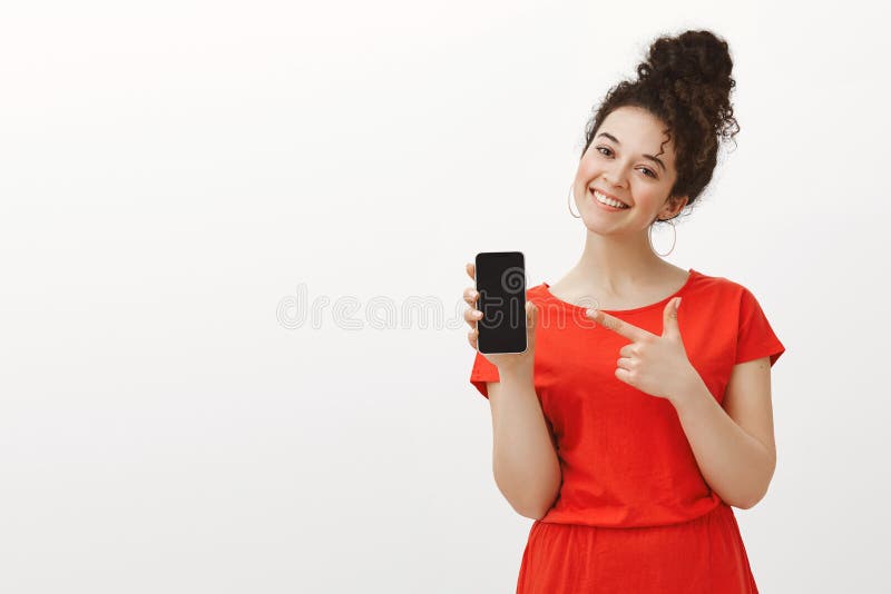 逗人喜爱的红色礼服的喜悦的无忧无虑的女朋友,显示智能手机和广泛地微笑与被掀动的头,当指向在时 库存照片 逗人喜爱的红色礼服的喜悦的无忧无虑的女朋友,显示智能手机和广泛地微笑与被掀动的头,当指向在时 库存照片