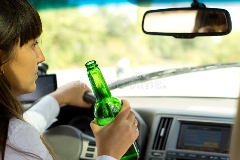 Closeup view from behind of a drunken female driver sitting steering the car while holding a bottle of booze and staring morosely ahead. Closeup view from behind of a drunken female driver sitting steering the car while holding a bottle of booze and staring morosely ahead