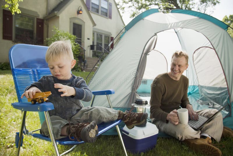 Father with laptop and son playing while camping in the front yard. Father with laptop and son playing while camping in the front yard