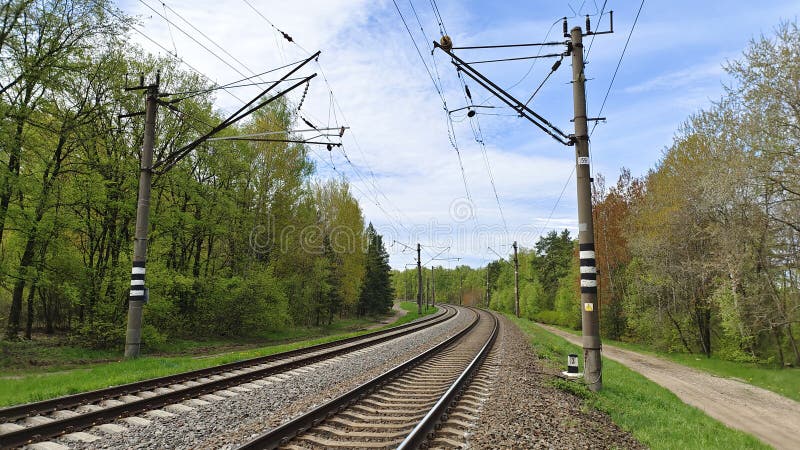 Railway rails are laid on concrete sleepers and sprinkled with crushed stone. The railroad is electrified, along the rails installed concrete poles with wires. Near there are grassy lawns, trees and country road. Railway rails are laid on concrete sleepers and sprinkled with crushed stone. The railroad is electrified, along the rails installed concrete poles with wires. Near there are grassy lawns, trees and country road