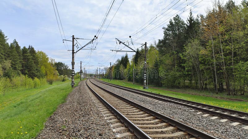 The railway rails are laid on reinforced concrete sleepers and sprinkled with rubble. The railway is electrified and there are concrete poles with wires along the rails Nearby there are grassy lawns and trees. The railway rails are laid on reinforced concrete sleepers and sprinkled with rubble. The railway is electrified and there are concrete poles with wires along the rails Nearby there are grassy lawns and trees