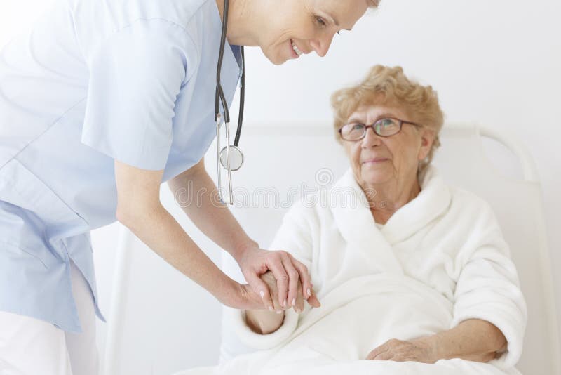 Smiling young doctor holding hand of elderly patient in white robe lying in bed. Smiling young doctor holding hand of elderly patient in white robe lying in bed