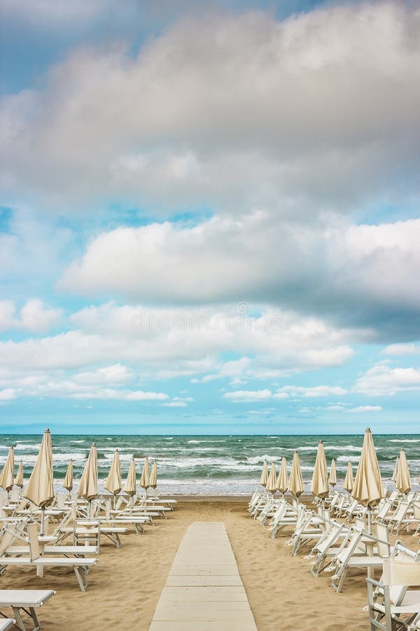 Rows of closed white umbrellas and deckchairs on the empty beach before the storm. The beginning or back-end of the season concept. Rows of closed white umbrellas and deckchairs on the empty beach before the storm. The beginning or back-end of the season concept.