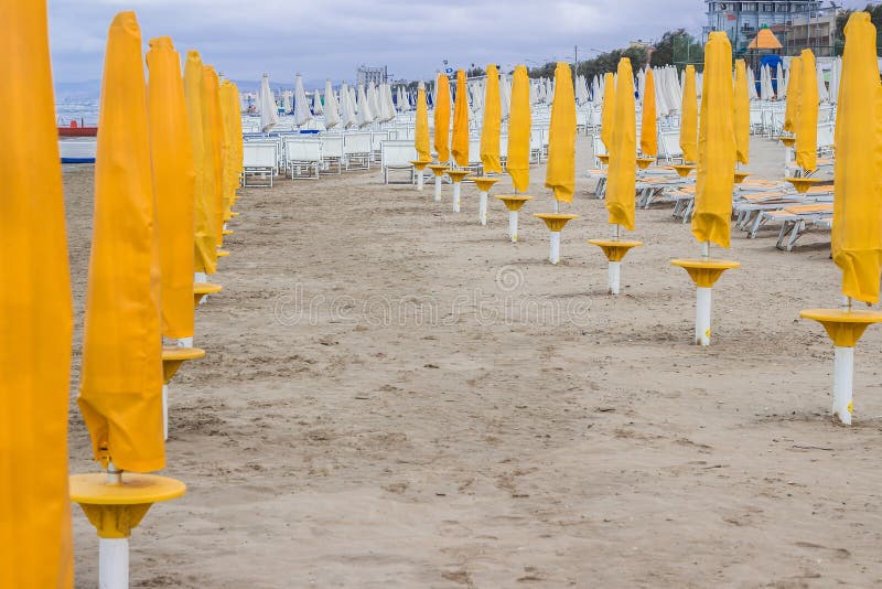 Rows of closed orange umbrellas and deckchairs on the empty beach before the storm. The beginning or back-end of the season concept. Rows of closed orange umbrellas and deckchairs on the empty beach before the storm. The beginning or back-end of the season concept.