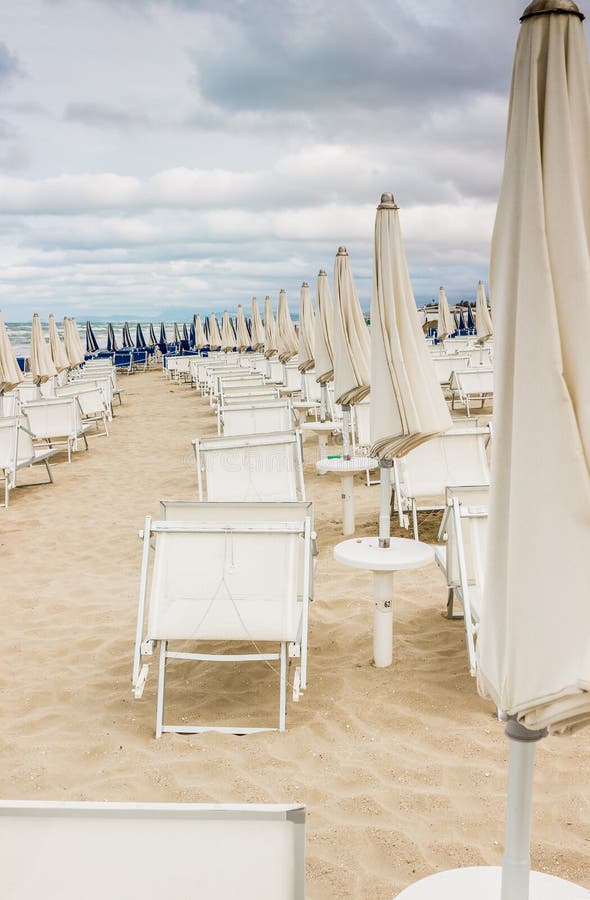 Rows of closed white umbrellas and deckchairs on the empty beach before the storm. The beginning or back-end of the season concept. Rows of closed white umbrellas and deckchairs on the empty beach before the storm. The beginning or back-end of the season concept.