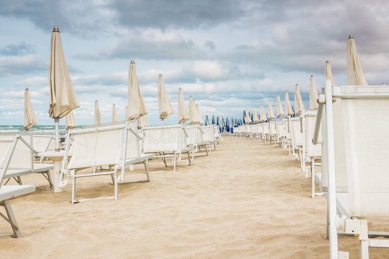 Rows of closed white umbrellas and deckchairs on the empty beach before the storm. The beginning or back-end of the season concept. Rows of closed white umbrellas and deckchairs on the empty beach before the storm. The beginning or back-end of the season concept.