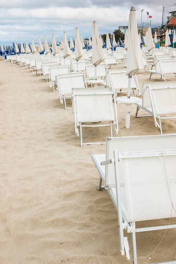 Rows of closed white umbrellas and deckchairs on the empty beach before the storm. The beginning or back-end of the season concept. Rows of closed white umbrellas and deckchairs on the empty beach before the storm. The beginning or back-end of the season concept.