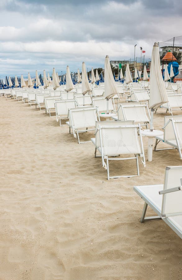 Rows of closed white umbrellas and deckchairs on the empty beach before the storm. The beginning or back-end of the season concept. Rows of closed white umbrellas and deckchairs on the empty beach before the storm. The beginning or back-end of the season concept.