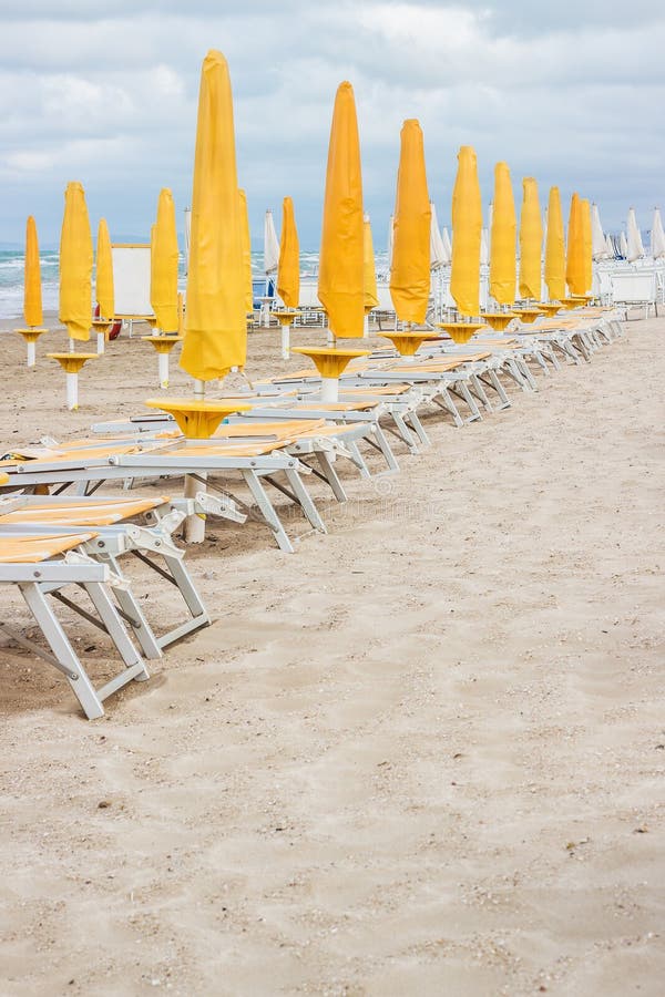 Rows of closed orange umbrellas and deckchairs on the empty beach before the storm. The beginning or back-end of the season concept. Rows of closed orange umbrellas and deckchairs on the empty beach before the storm. The beginning or back-end of the season concept.