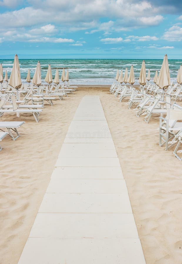 Rows of closed white umbrellas and deckchairs on the empty beach before the storm. The beginning or back-end of the season concept. Rows of closed white umbrellas and deckchairs on the empty beach before the storm. The beginning or back-end of the season concept.