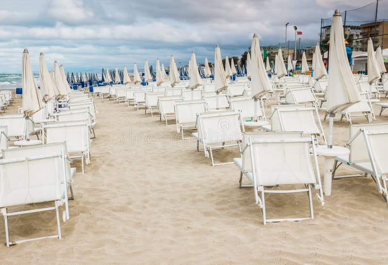 Rows of closed white umbrellas and deckchairs on the empty beach before the storm. The beginning or back-end of the season concept. Rows of closed white umbrellas and deckchairs on the empty beach before the storm. The beginning or back-end of the season concept.