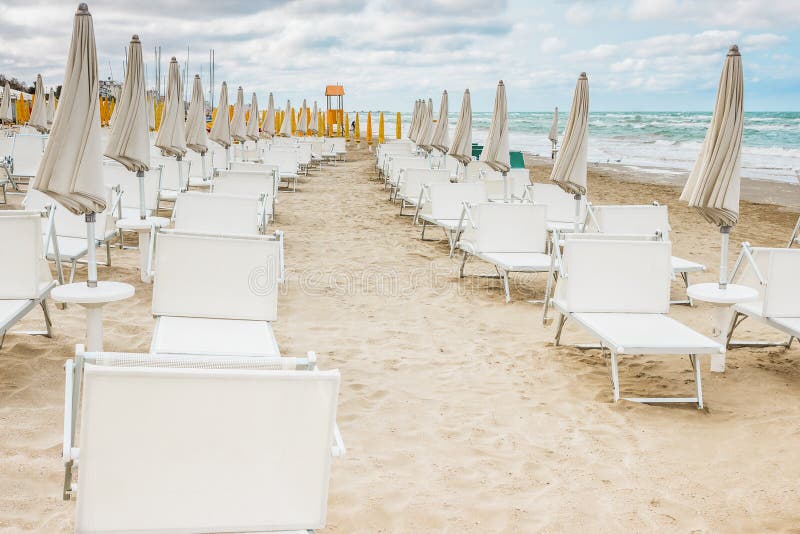 Rows of closed white umbrellas and deckchairs on the empty beach before the storm. The beginning or back-end of the season concept. Rows of closed white umbrellas and deckchairs on the empty beach before the storm. The beginning or back-end of the season concept.