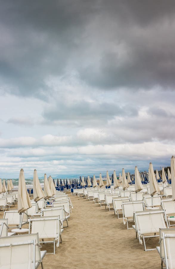 Rows of closed white umbrellas and deckchairs on the empty beach before the storm. The beginning or back-end of the season concept. Rows of closed white umbrellas and deckchairs on the empty beach before the storm. The beginning or back-end of the season concept.