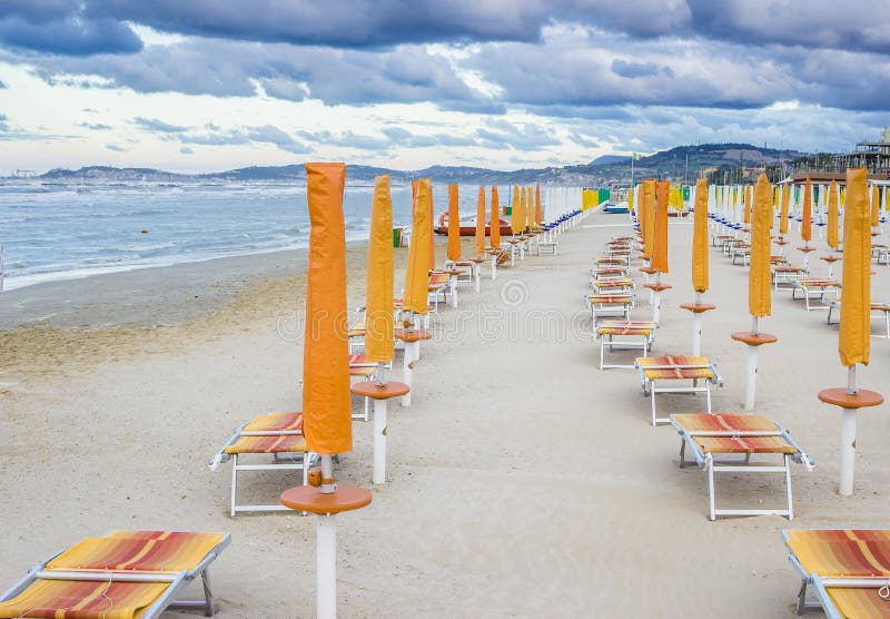 Rows of closed orange umbrellas and deckchairs on the empty beach before the storm. The beginning or back-end of the season concept. Rows of closed orange umbrellas and deckchairs on the empty beach before the storm. The beginning or back-end of the season concept.