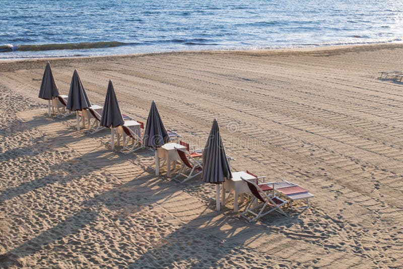 Line of closed beach umbrellas and empty loungers, chairs and sunbeds, with no people. Line of closed beach umbrellas and empty loungers, chairs and sunbeds, with no people