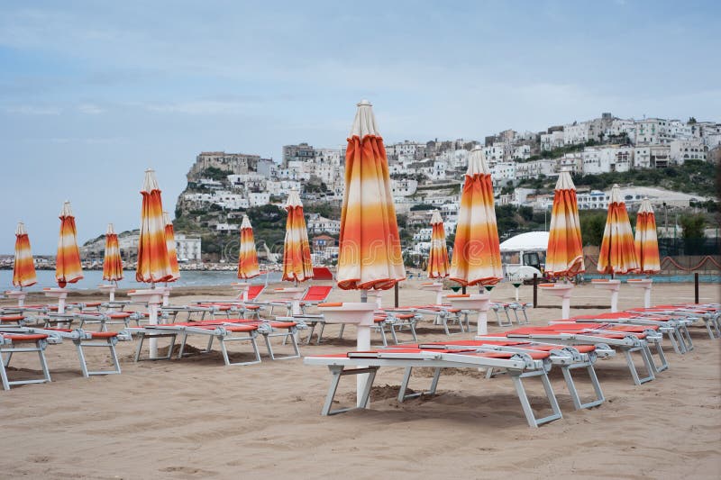 Closed orange beach umbrellas and deck chairs on the beach of Peschici, Apulia, Italy on a cold spring day. Closed orange beach umbrellas and deck chairs on the beach of Peschici, Apulia, Italy on a cold spring day.