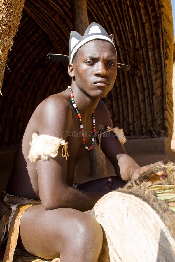 An african zulu tribe young man wearing traditional clothes sitting inside a hut and playing with an african drum. An african zulu tribe young man wearing traditional clothes sitting inside a hut and playing with an african drum