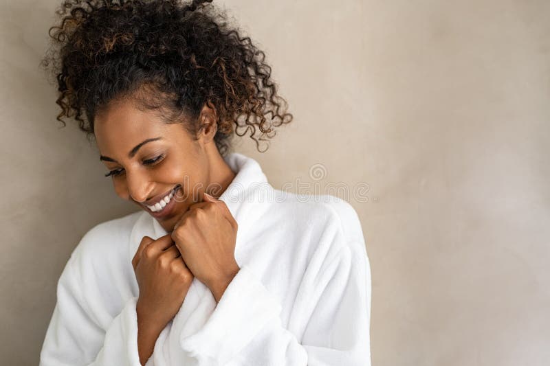 Beautiful young woman in a white bath robe  on beige background with copy space. Attractive african girl in bathrobe looking down against light brown wall. Happy african american woman enjoy after body treatment. Beautiful young woman in a white bath robe  on beige background with copy space. Attractive african girl in bathrobe looking down against light brown wall. Happy african american woman enjoy after body treatment